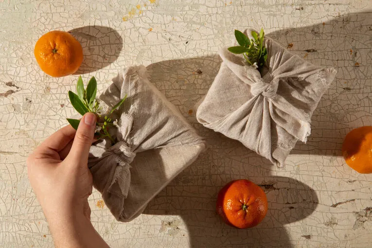 A hand adjusting a sustainably wrapped gift box adorned with greenery. Other eco-friendly gift packages and tangerines are visible.