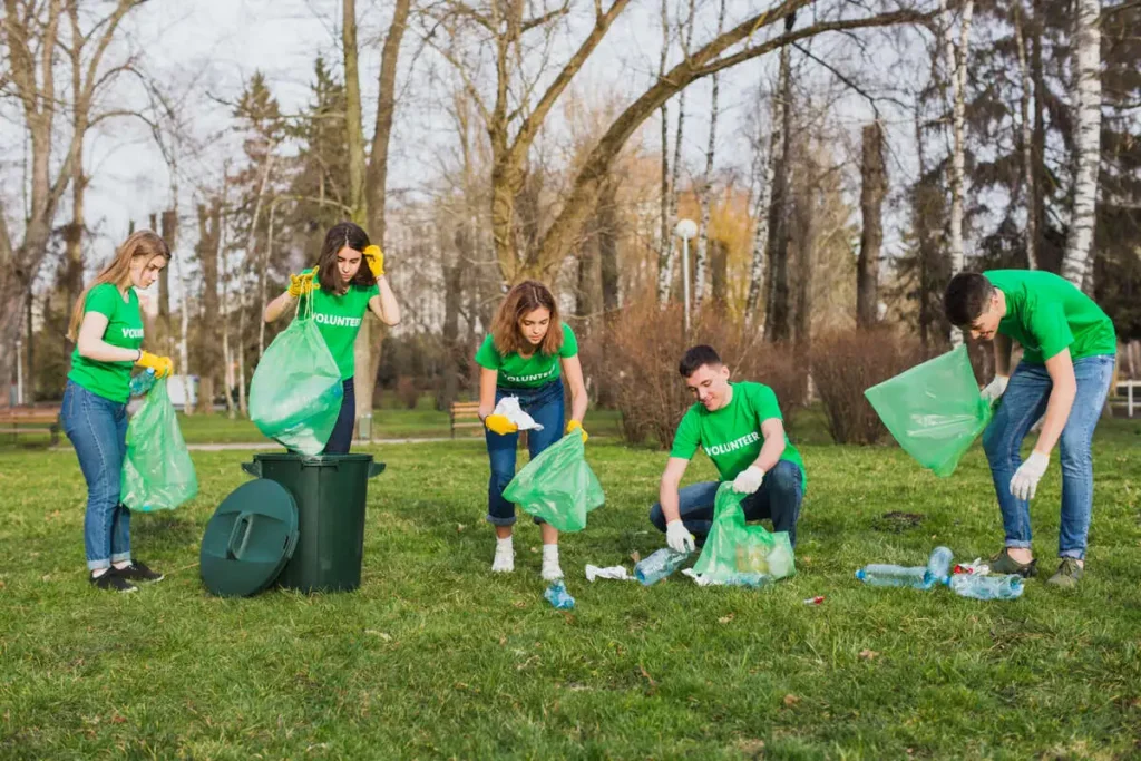 A group of volunteers engaged in an environment cleanliness drive are cleaning up a park by picking up litter and placing it into green trash bags. The park is filled with trees and grass.